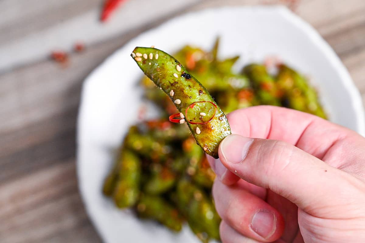 Stir fried edamame with garlic and oyster sauce glaze topped with dried chili and sesame seeds on a white plate