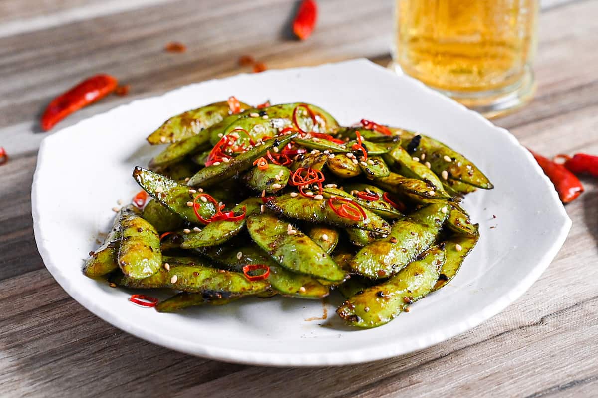 Stir fried edamame with garlic and oyster sauce glaze topped with dried chili and sesame seeds on a white plate side