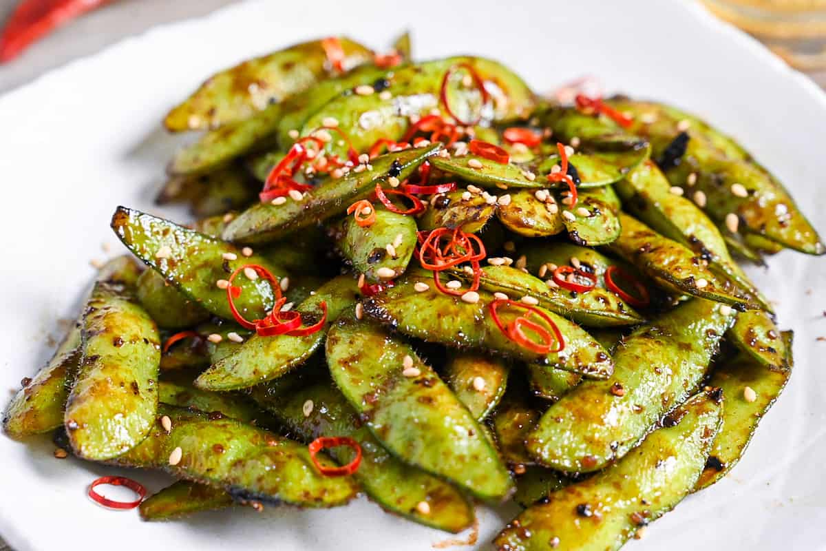Stir fried edamame with garlic and oyster sauce glaze topped with dried chili and sesame seeds on a white plate close up