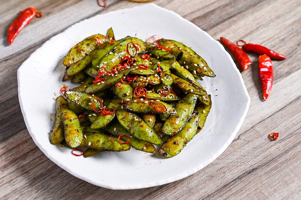 Stir fried edamame with garlic and oyster sauce glaze topped with dried chili and sesame seeds on a white plate