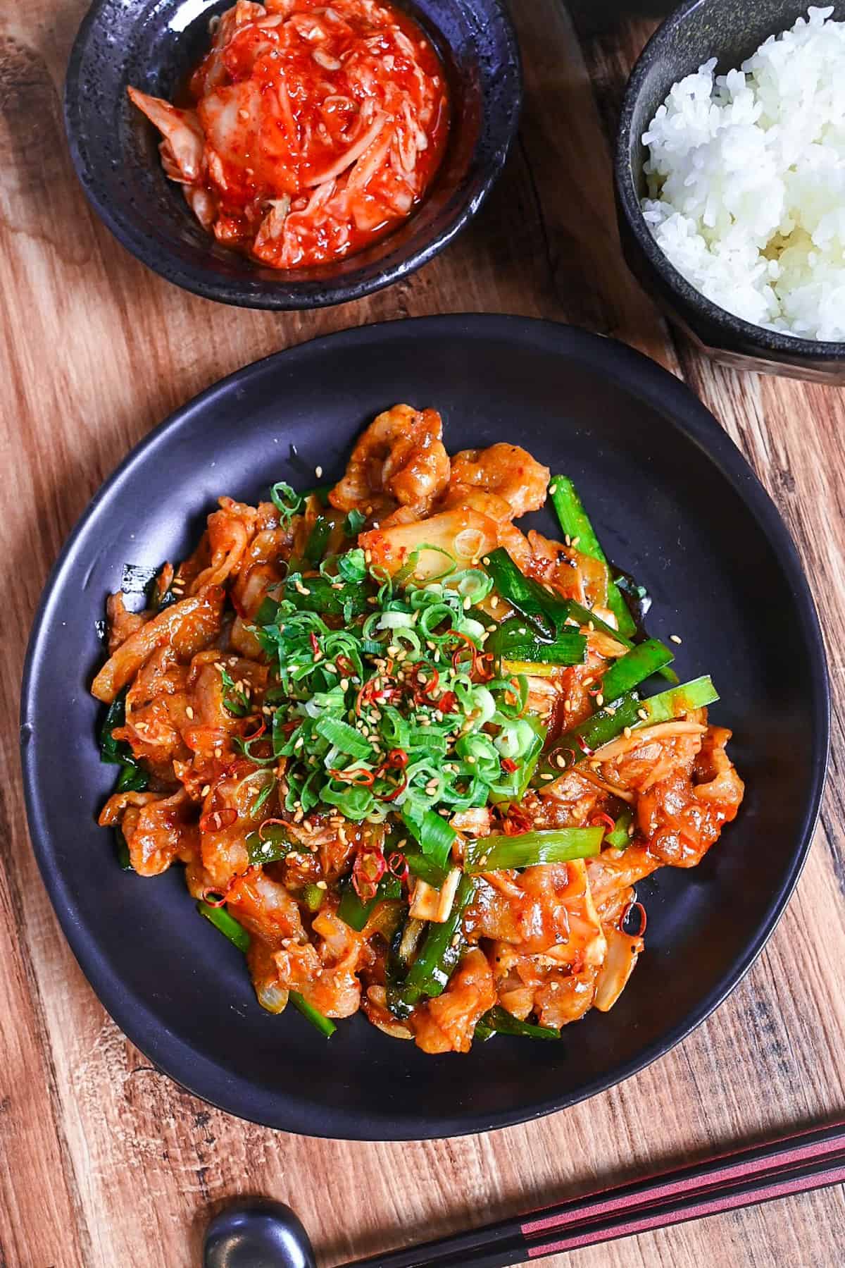 Japanese buta kimchi itame (pork and kimchi stir fry) on a black plate topped with chopped green onion, dried chili and sesame seeds served next to a bowl of kimchi, rice and miso soup.