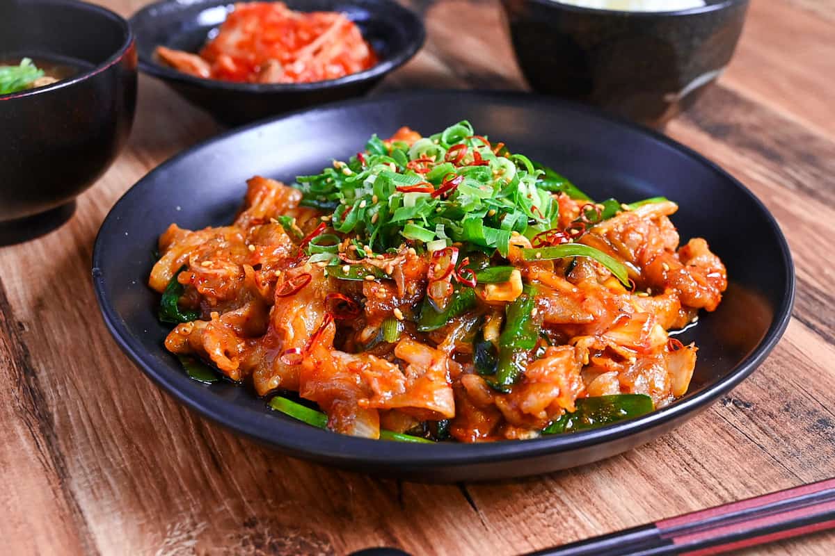 Japanese buta kimchi itame (pork and kimchi stir fry) on a black plate topped with chopped green onion, dried chili and sesame seeds served next to a bowl of kimchi, rice and miso soup. (side view)