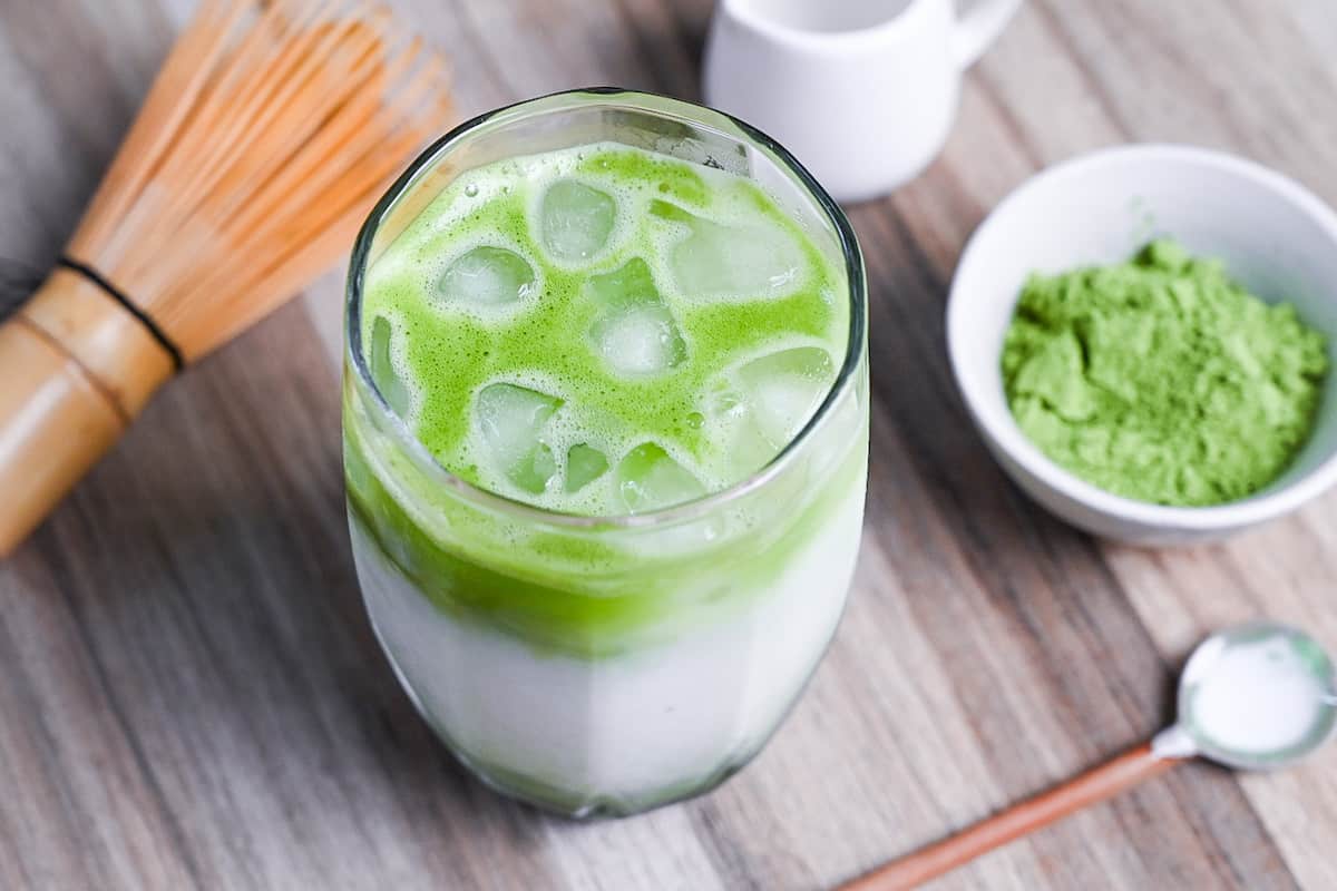 iced matcha latte with matcha whisk and small white bowl of matcha powder