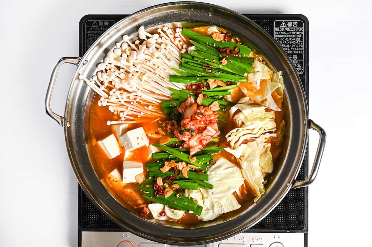 kimchi nabe with firm tofu, enoki mushrooms, pork belly and cabbage topped with garlic chives, fried garlic, sesame seeds, chili and kimchi