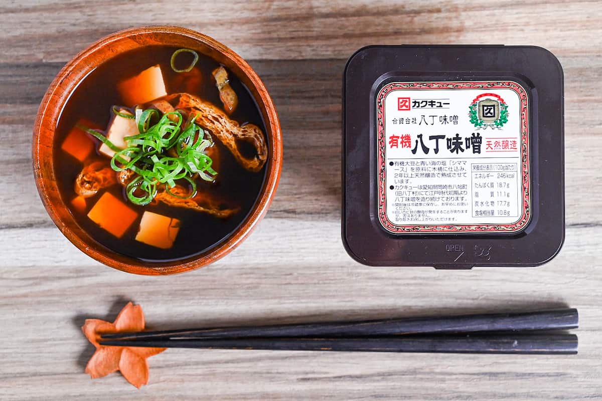 aka-dashi (red miso soup) in wooden bowl topped with chopped green onion next to a box of hatcho red miso