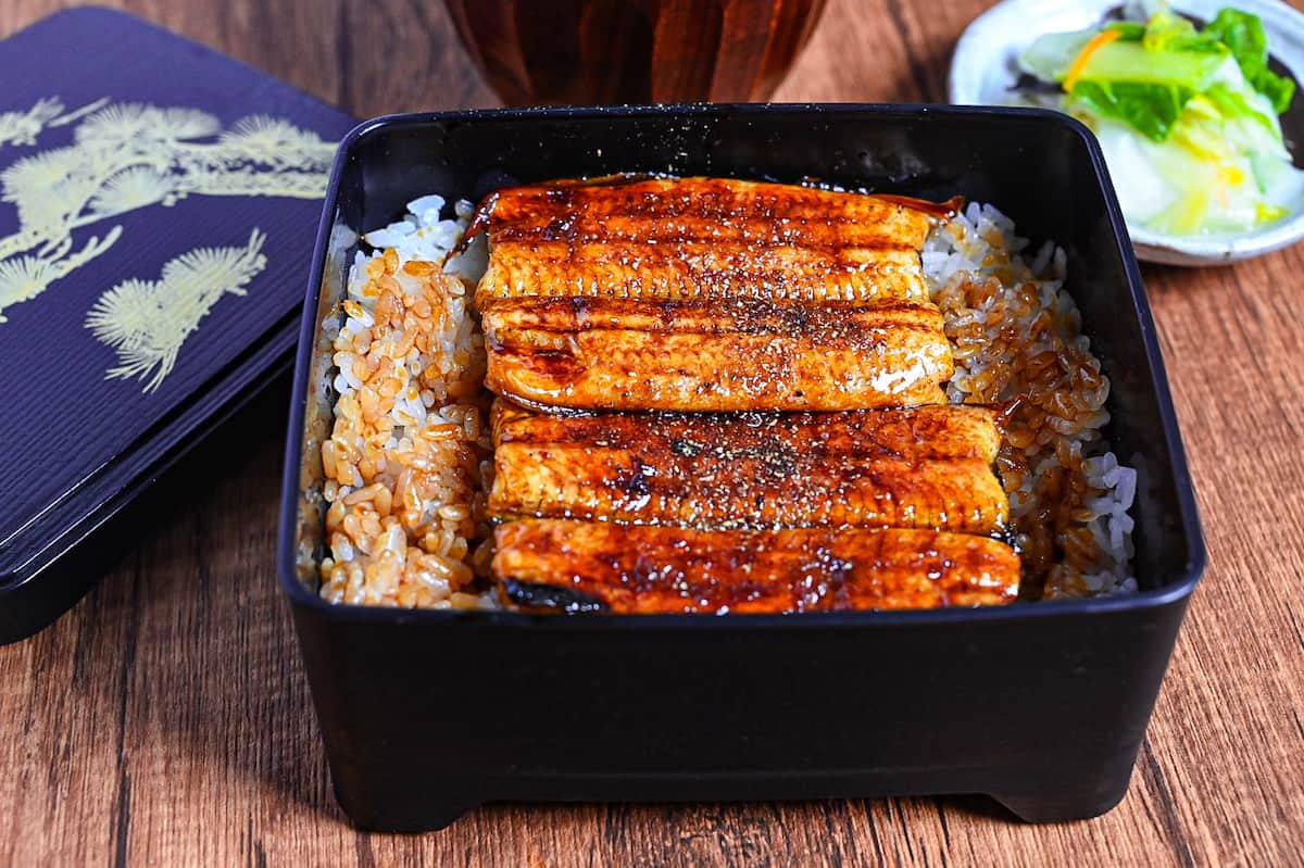 grilled eel coated with homemade unagi sauce and sprinkled with sansho pepper served over rice in a black lacquerware box (unaju) with a plate of pickles and bowl of clear soup