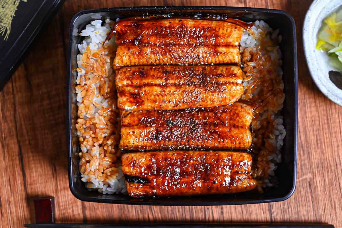 grilled eel coated with homemade unagi sauce and sprinkled with sansho pepper served over rice in a black lacquerware box (unaju) with a plate of pickles and bowl of clear soup