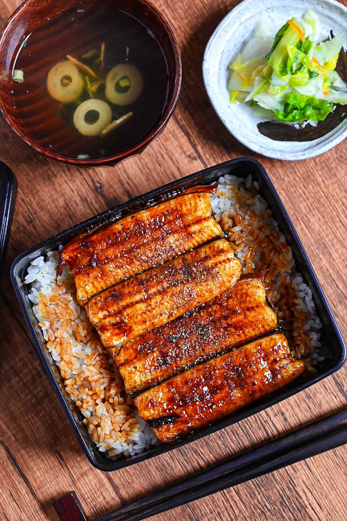 grilled eel coated with homemade unagi sauce and sprinkled with sansho pepper served over rice in a black lacquerware box (unaju) with a plate of pickles and bowl of clear soup