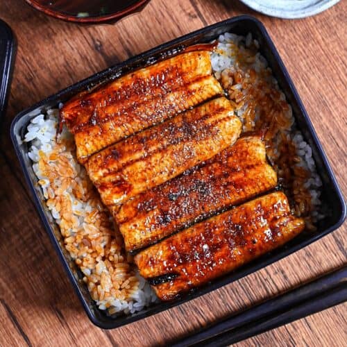 grilled eel coated with homemade unagi sauce and sprinkled with sansho pepper served over rice in a black lacquerware box (unaju) with a plate of pickles and bowl of clear soup