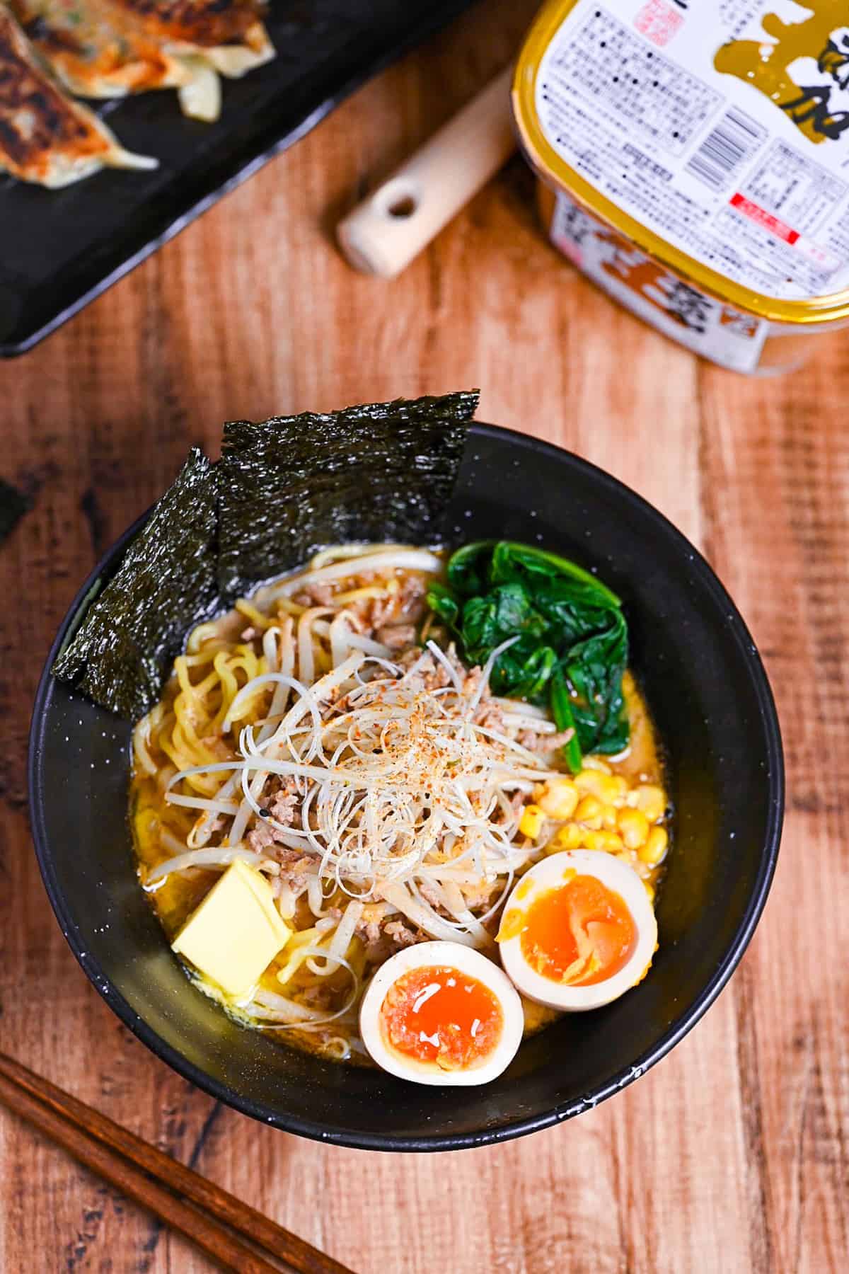 Easy homemade pork miso ramen in a black bowl topped with ramen eggs, spinach, beansprouts, nori and sweetcorn next to a plate of fried gyoza and a tub of miso paste.