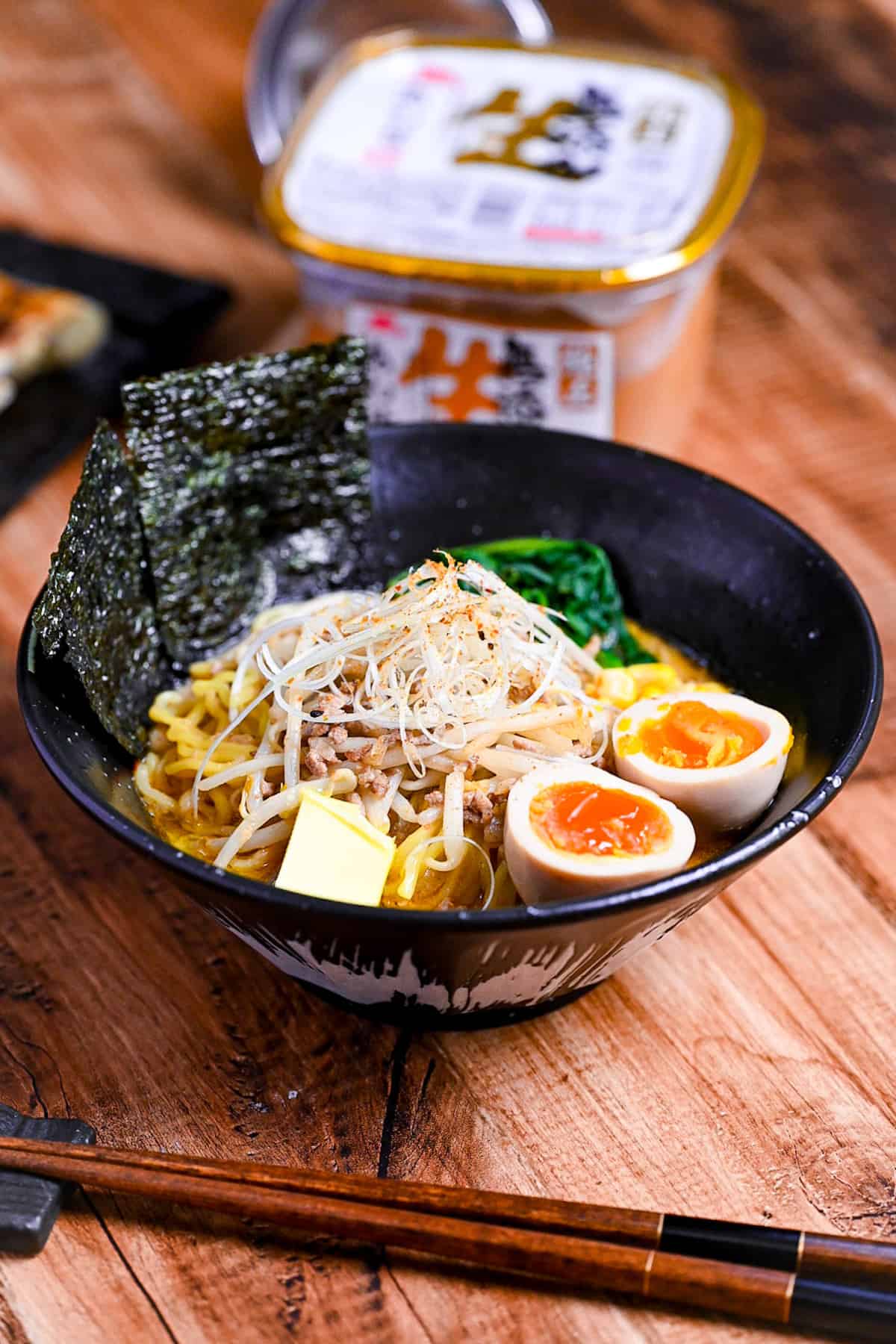 Easy homemade pork miso ramen in a black bowl topped with ramen eggs, spinach, beansprouts, nori and sweetcorn next to a plate of fried gyoza and a tub of miso paste.