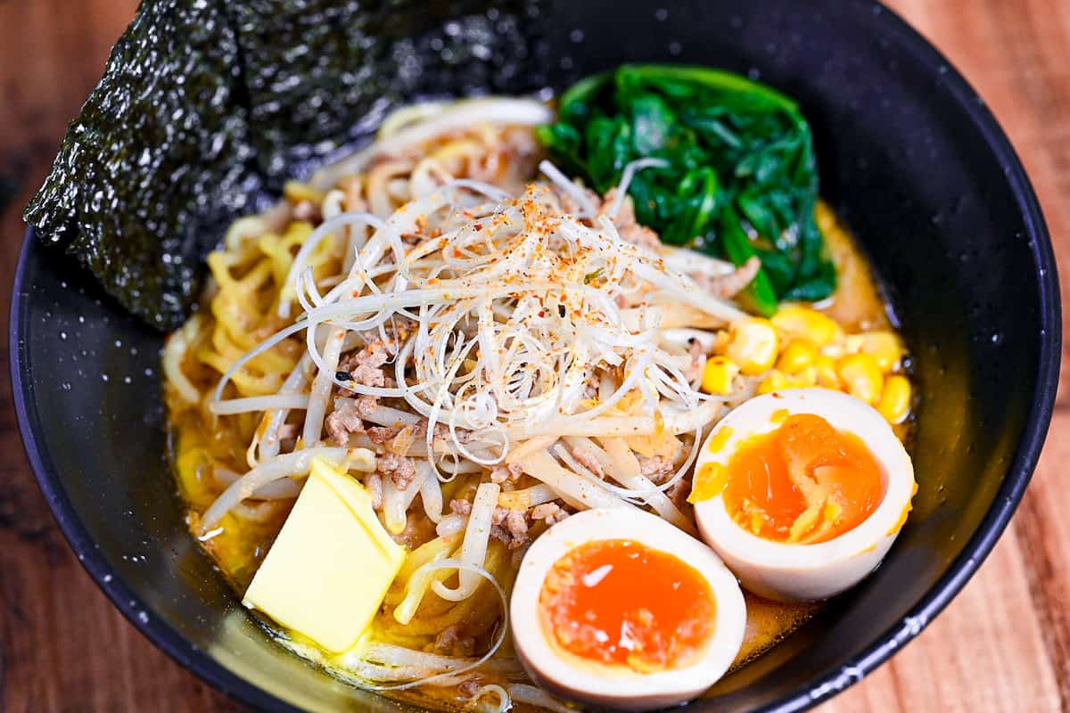 Easy homemade pork miso ramen in a black bowl topped with ramen eggs, spinach, beansprouts, nori and sweetcorn next to a plate of fried gyoza and a tub of miso paste.