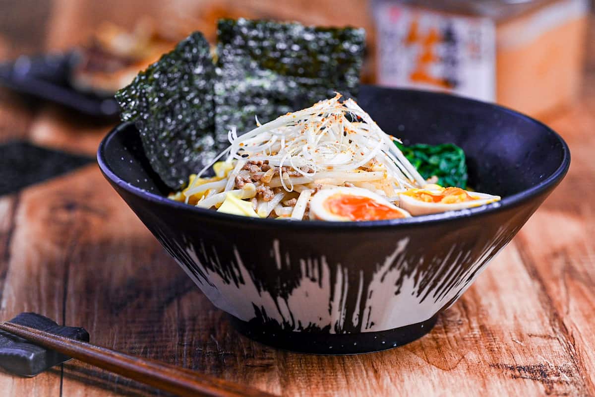 Easy homemade pork miso ramen in a black bowl topped with ramen eggs, spinach, beansprouts, nori and sweetcorn next to a plate of fried gyoza and a tub of miso paste.