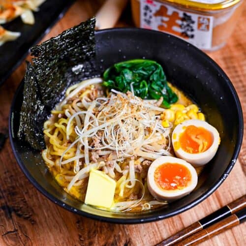 Easy homemade pork miso ramen in a black bowl topped with ramen eggs, spinach, beansprouts, nori and sweetcorn next to a plate of fried gyoza and a tub of miso paste.