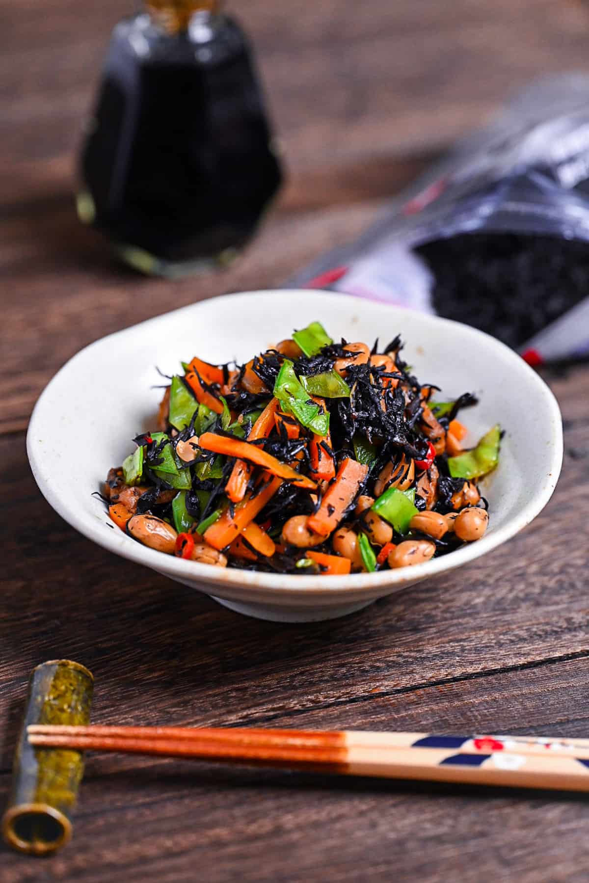 hijiki salad in a white dish next to a bottle of soy sauce, dried hijiki and wooden chopsticks on a bamboo shaped rest