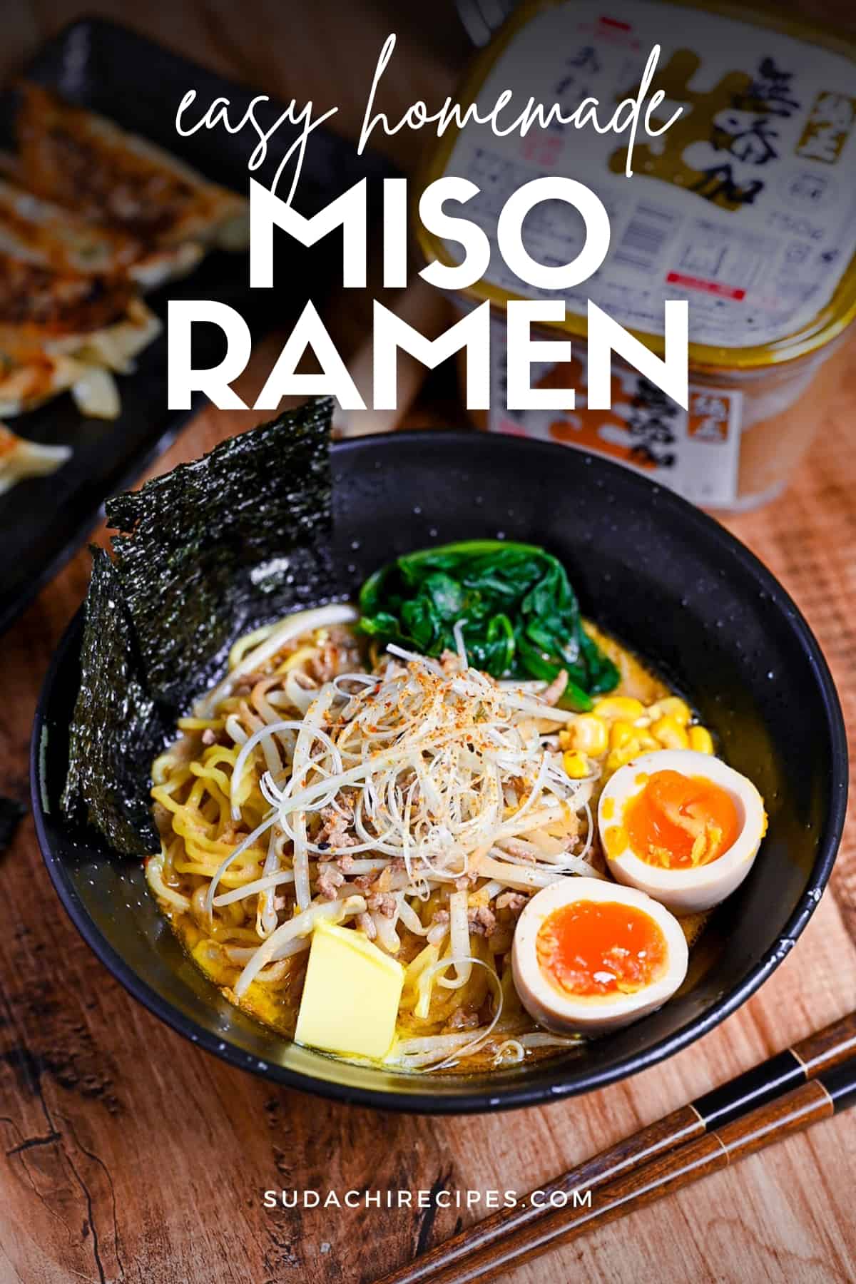 Easy homemade pork miso ramen in a black bowl topped with ramen eggs, spinach, beansprouts, nori and sweetcorn next to a plate of fried gyoza and a tub of miso paste.