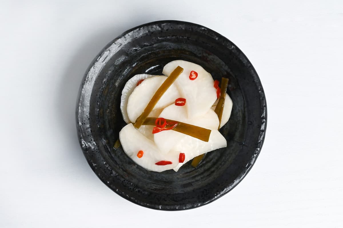 bettarazuke sweet pickled daikon radish on a black plate topped with thin slices of kombu and dried red chili