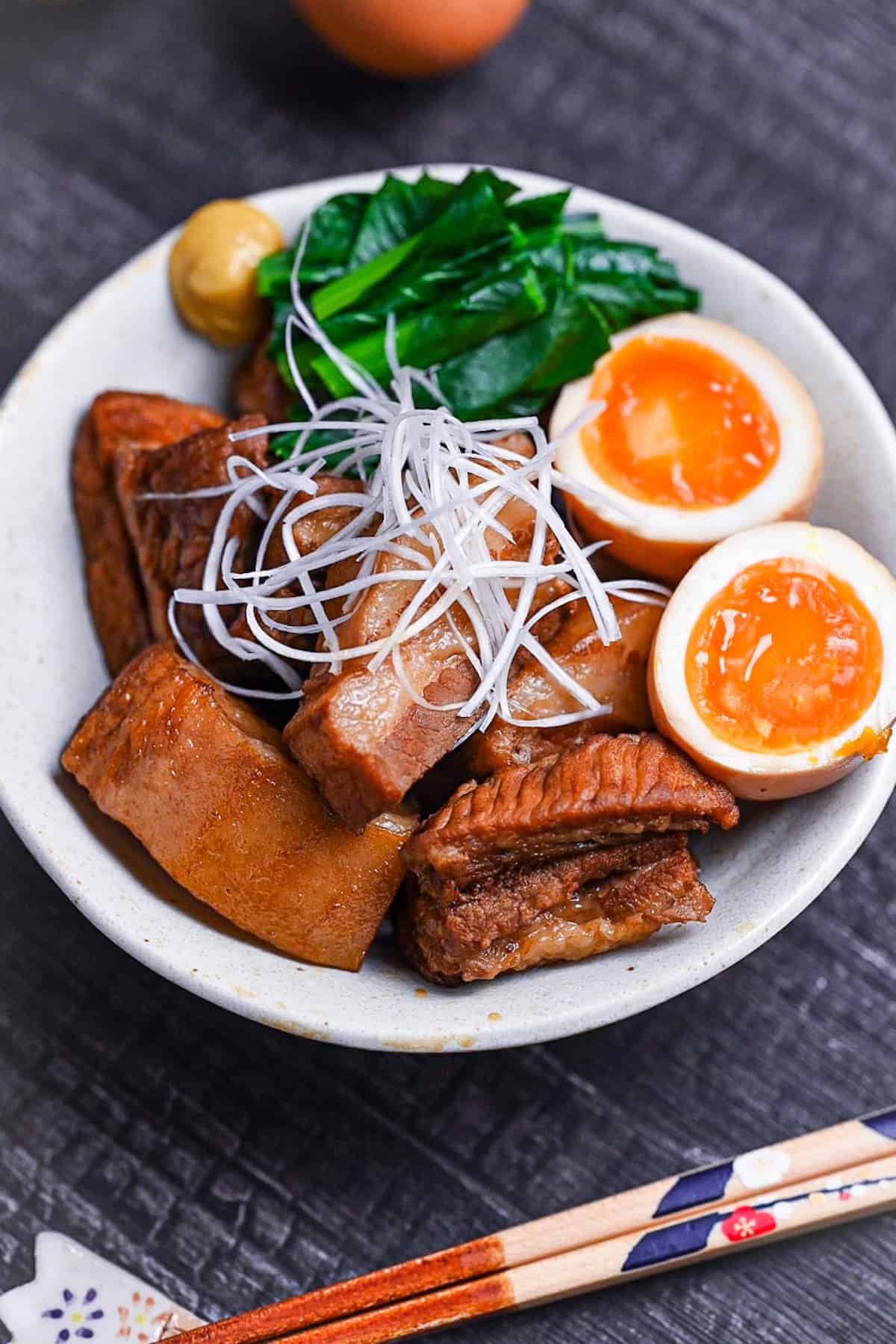 Buta no kakuni (Japanese braised pork belly) in an off-white dish with marinated soft boiled eggs, mustard spinach and a blob of mustard topped with shredded green onion