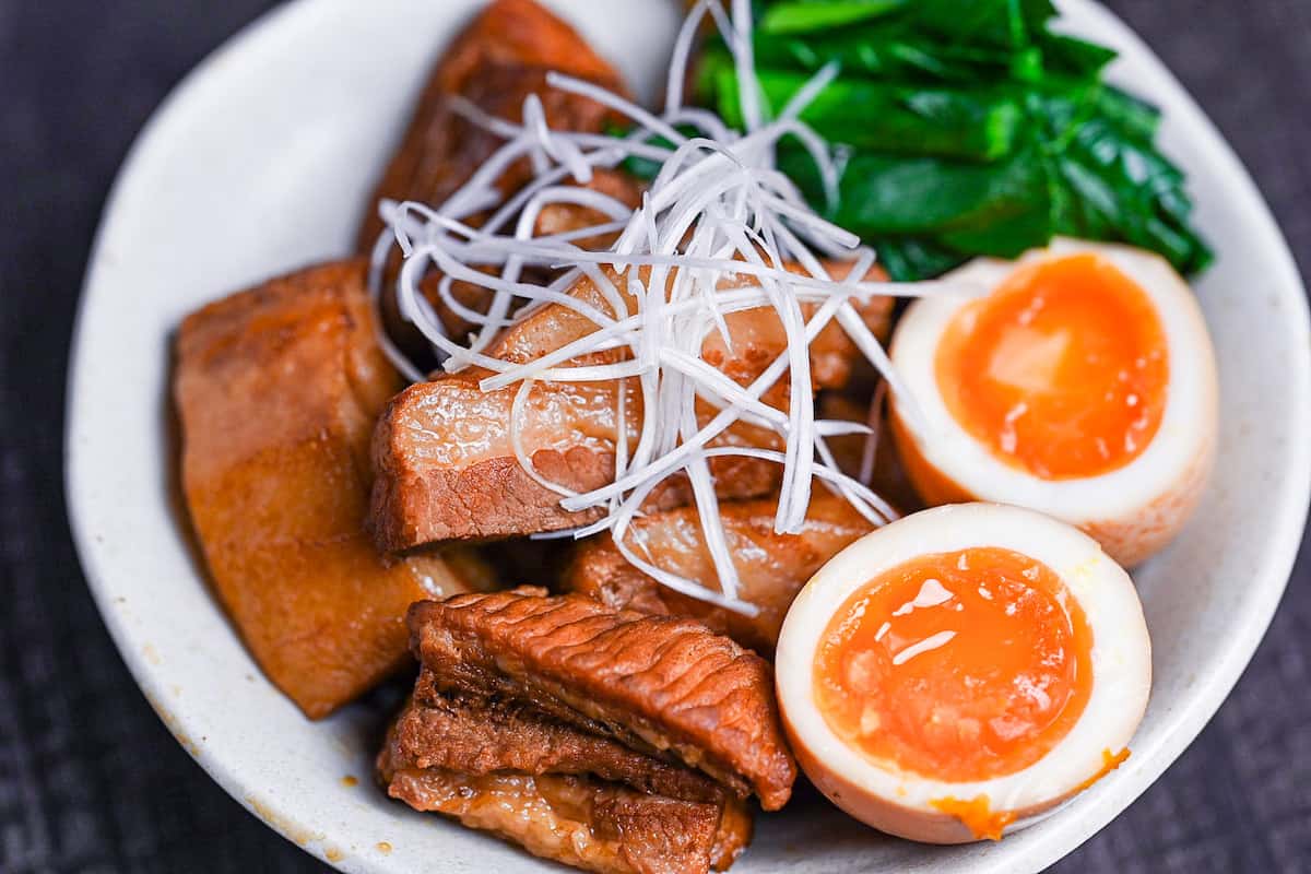 Buta no kakuni (Japanese braised pork belly) in an off-white dish with marinated soft boiled eggs, mustard spinach and a blob of mustard topped with shredded green onion