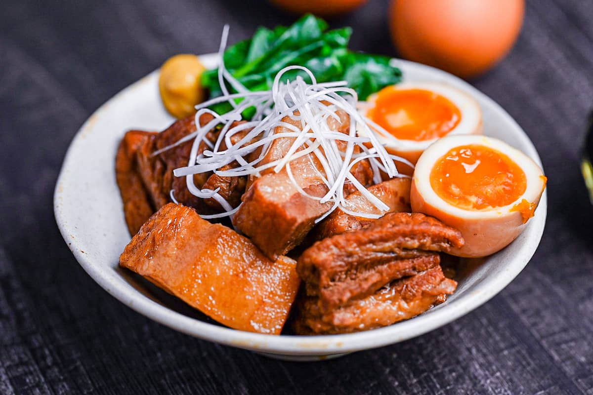 Buta no kakuni (Japanese braised pork belly) in an off-white dish with marinated soft boiled eggs, mustard spinach and a blob of mustard topped with shredded green onion