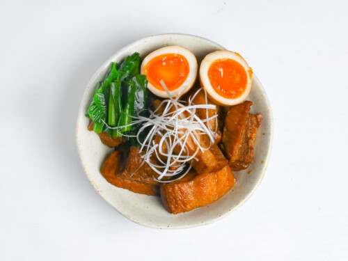 buta no kakuni in an off-white dish next to soft boiled eggs and greens topped with shiraganegi