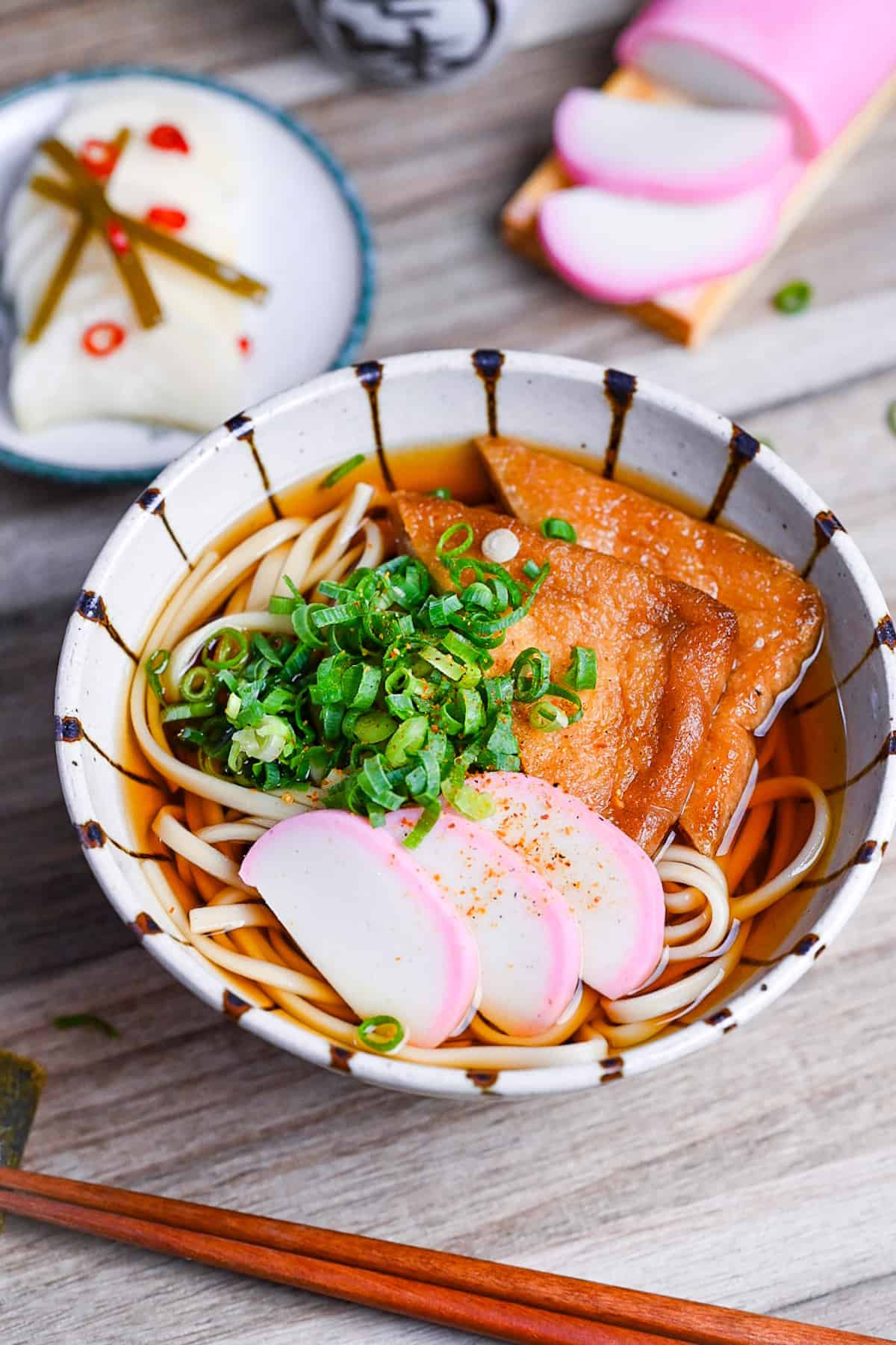 Kitsune Udon made with udon noodles in a dashi soup topped with fried tofu pouches, pink and white fish cakes (kamaboko) and chopped green onions in a beige bowl with brown stripes