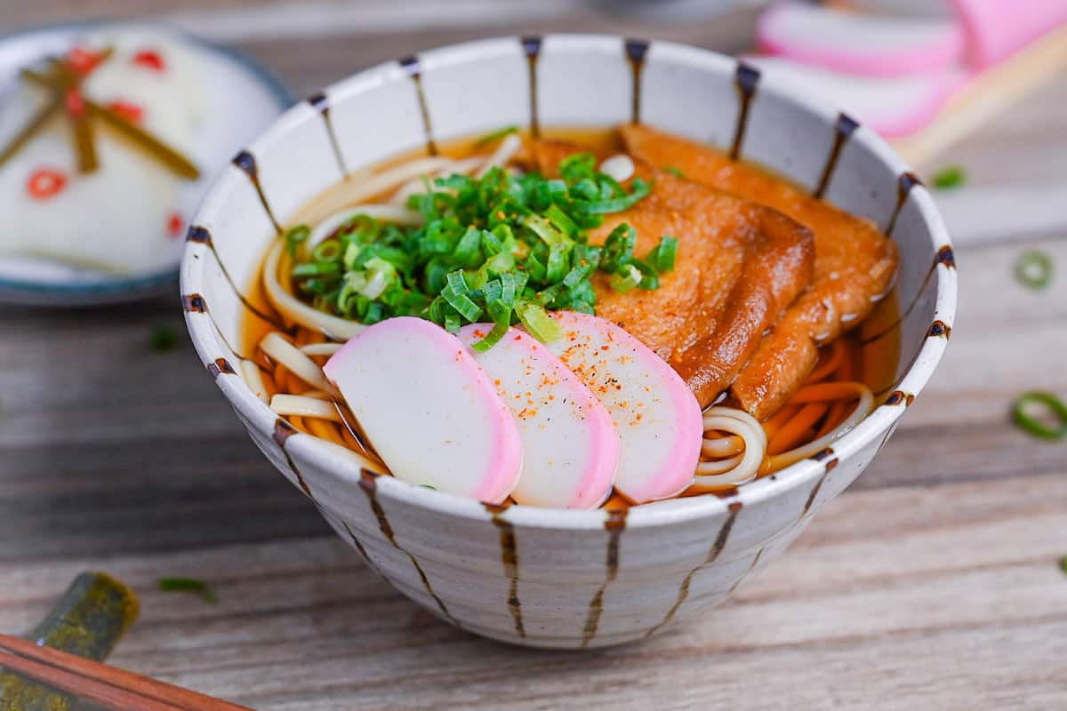 Kitsune Udon made with udon noodles in a dashi soup topped with fried tofu pouches, pink and white fish cakes (kamaboko) and chopped green onions in a beige bowl with brown stripes