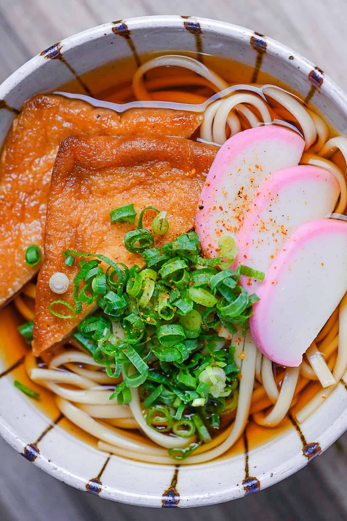 Kitsune Udon made with udon noodles in a dashi soup topped with fried tofu pouches, pink and white fish cakes (kamaboko) and chopped green onions in a beige bowl with brown stripes