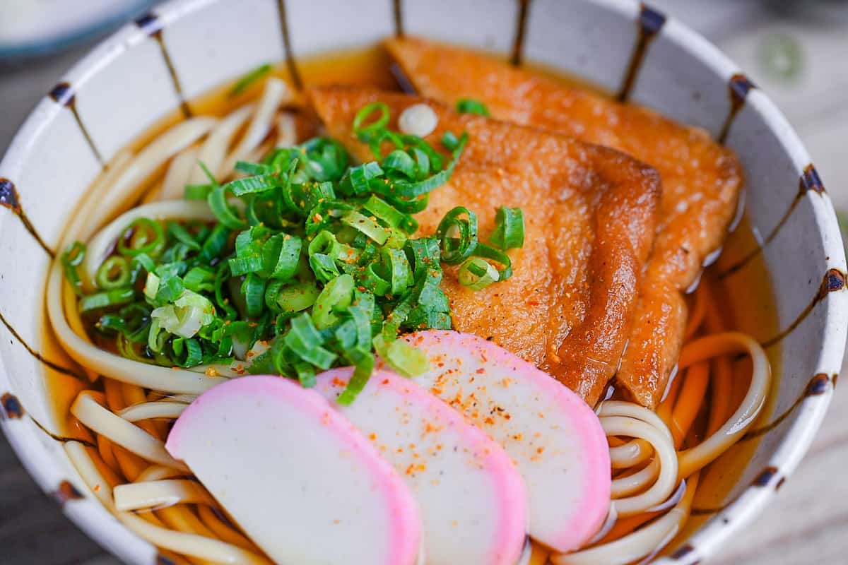 Kitsune Udon made with udon noodles in a dashi soup topped with fried tofu pouches, pink and white fish cakes (kamaboko) and chopped green onions in a beige bowl with brown stripes