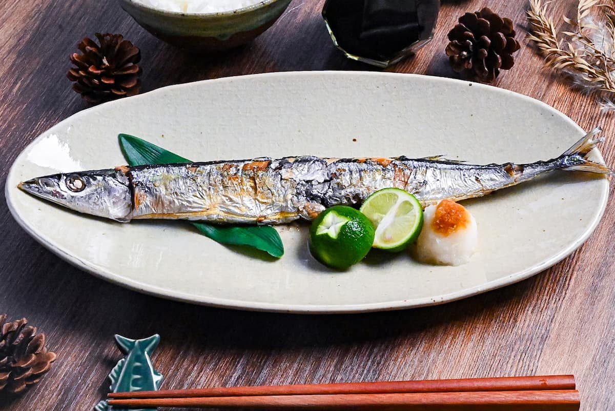 Grilled pacific saury (sanma no shioyaki) on a pale oval plate with grated daikon, sauce sauce and a halved sudachi
