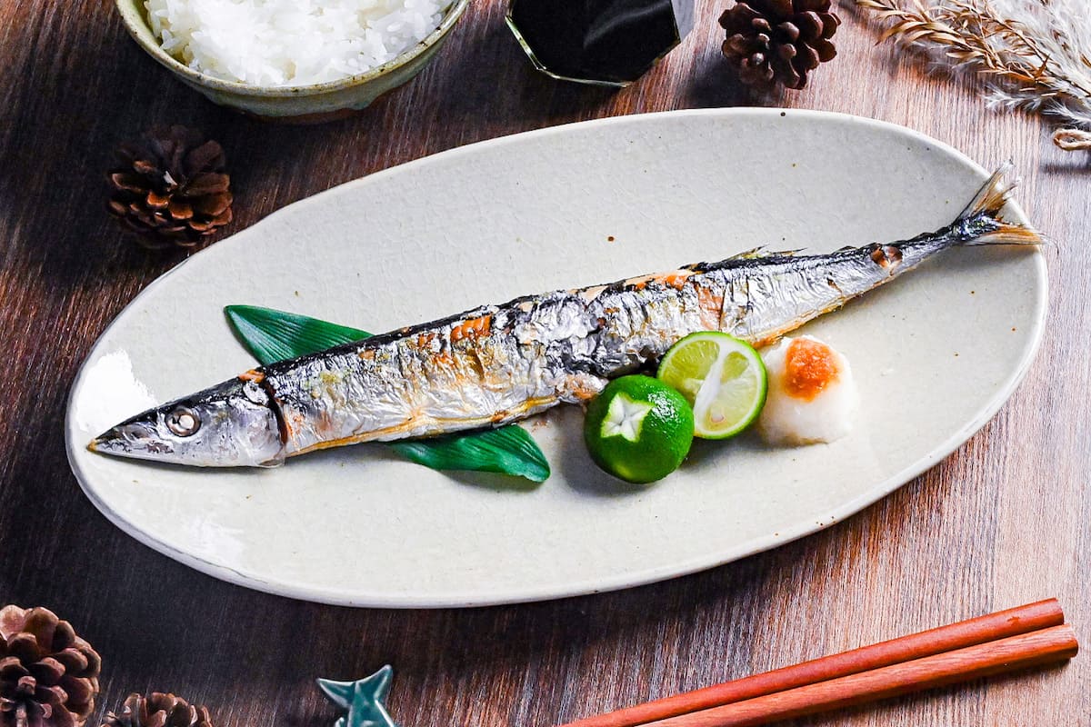 Grilled pacific saury (sanma no shioyaki) on a pale oval plate with grated daikon, sauce sauce and a halved sudachi