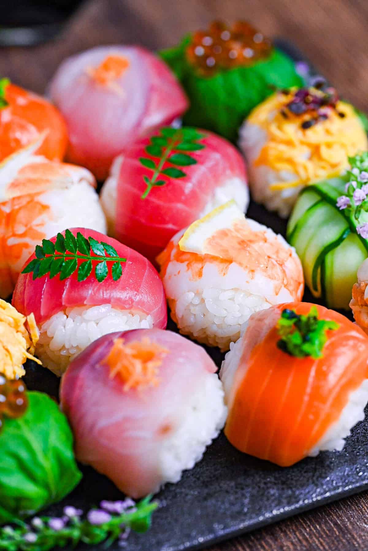 decorative temarizushi made with various seafood, eggs and vegetables topped with leaves, salmon roe, flowers and lemon slices served on a square slate-style plate