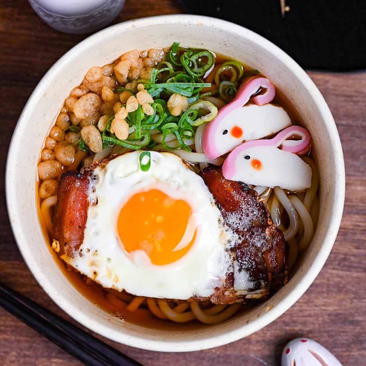 Tsukimi udon made with thick noodles in a dashi broth topped with eggs, bacon, green onion, tempura bits and kamaboko fishcake shaped into a rabbit