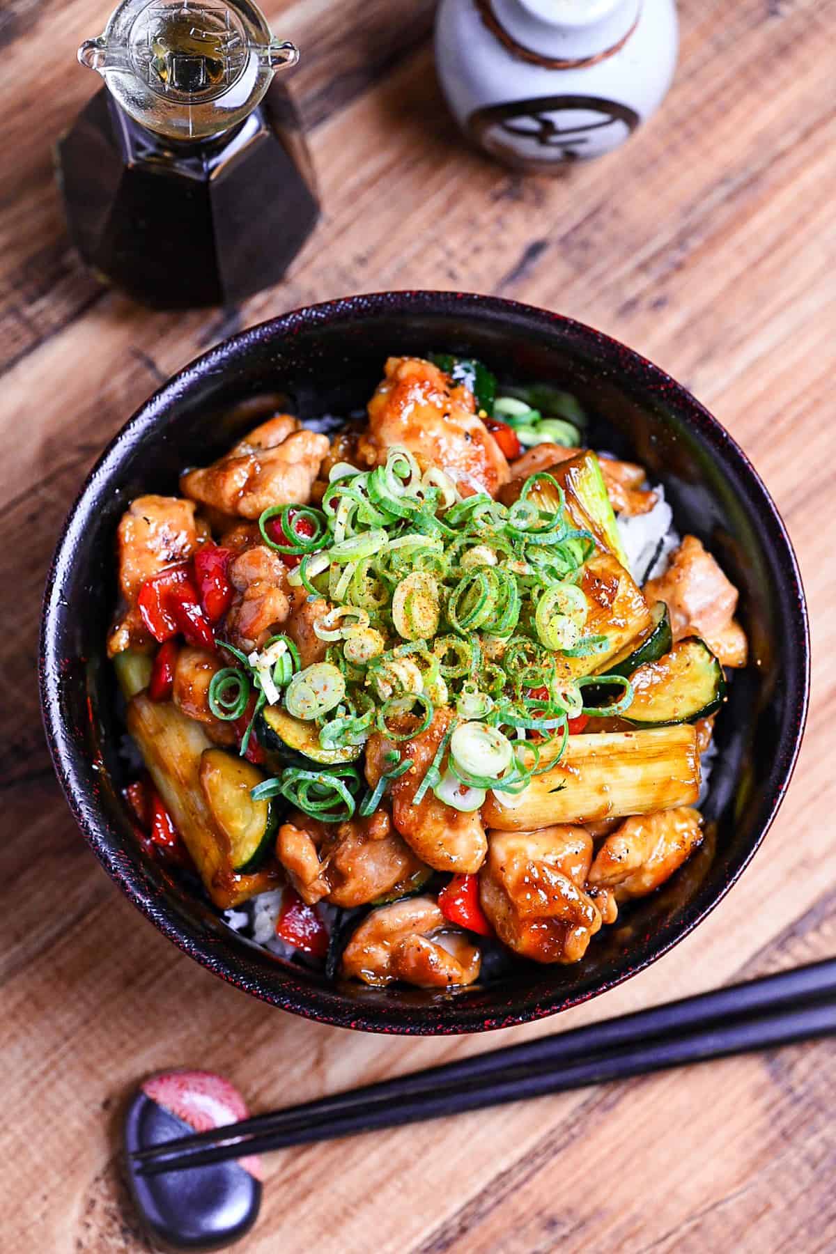Yakitori donburi made with chicken, zucchini, green onions and bell peppers cooked in a yakitori-style tare (sauce) served in a black bowl and topped with green onion and shichimi