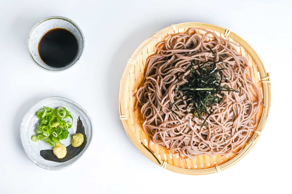 zaru soba on a bamboo tray topped with nori and served next to homemade mentsuyu dipping sauce and a small plate with chopped green onion, wasabi and grated ginger