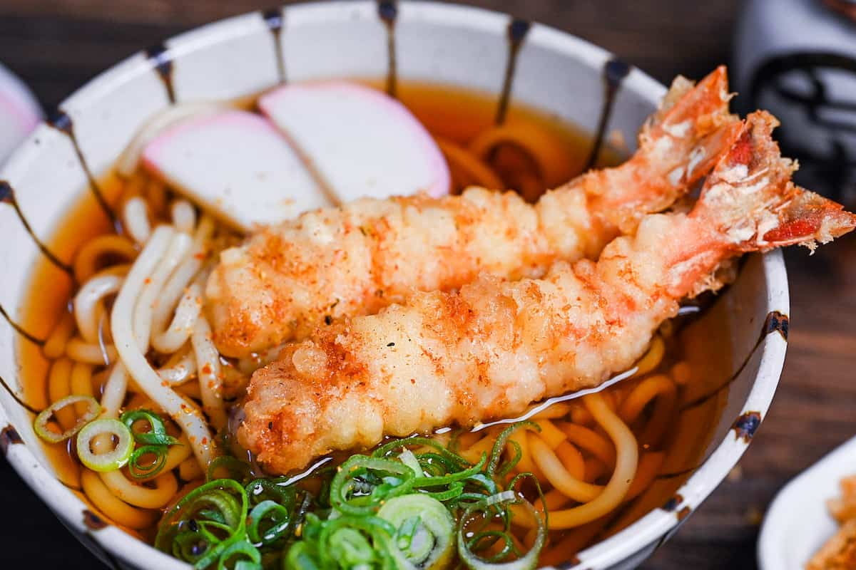 Ebiten Udon - Homemade udon noodle soup in a stripy bowl topped with kamaboko, green onion and tempura shrimp