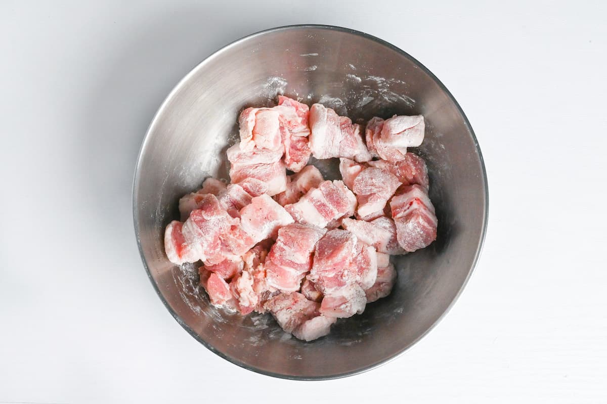 cubes of raw pork belly coated with a thin layer of flour in a steel mixing bowl