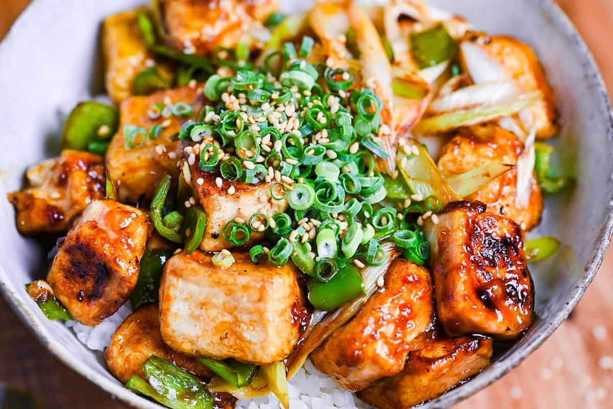 tofu donburi with teriyaki glaze topped with chili oil, green onion and sesame seeds over rice in a mottled bowl