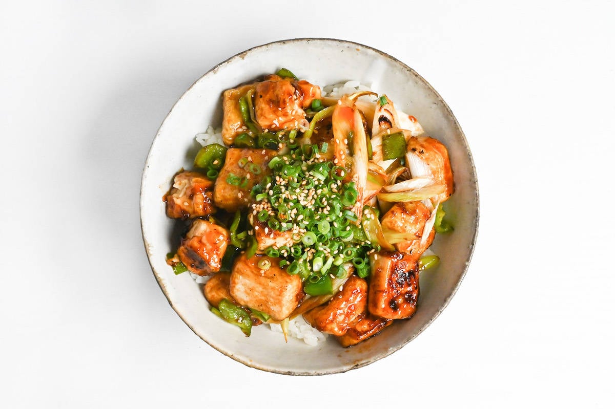 tofu donburi with teriyaki glaze topped with chili oil, green onion and sesame seeds over rice in a mottled bowl