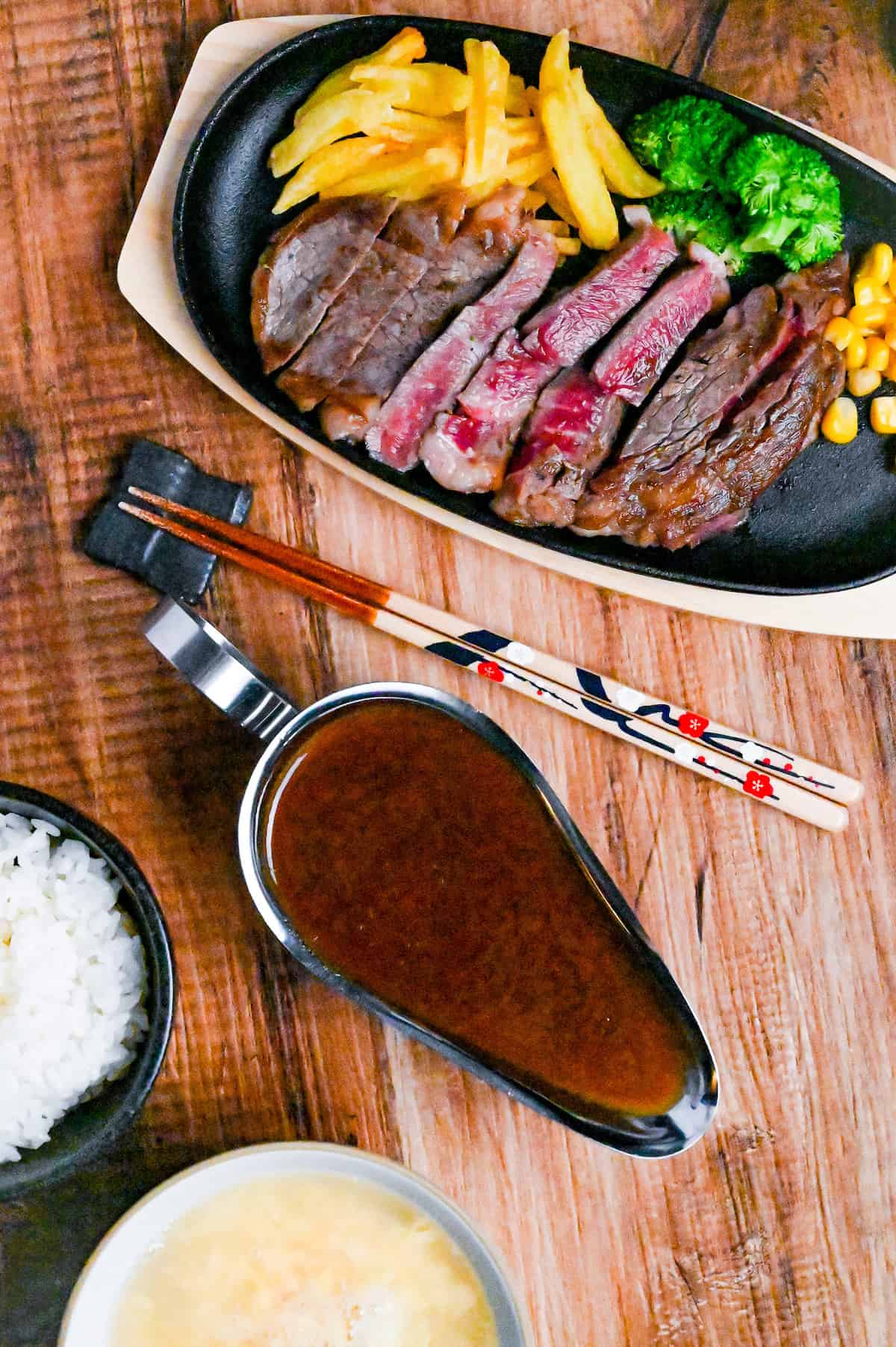 Japanese steakhouse style steak sauce in a steel gravy boat next to a iron plate with slices of rare beef steak, fries and vegetables top down view