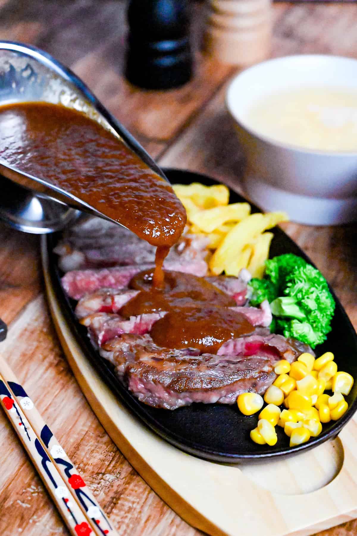 slices of rare steak drizzled with Japanese style steak sauce served on a black hotplate with fries, broccoli and corn next to a steel gravy boat filled with steak sauce