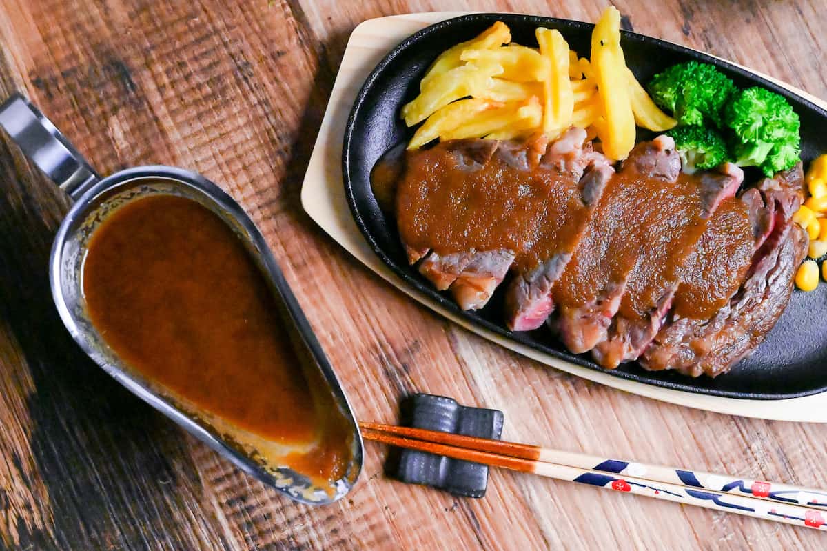 slices of rare steak drizzled with Japanese style steak sauce served on a black hotplate with fries, broccoli and corn next to a steel gravy boat filled with steak sauce