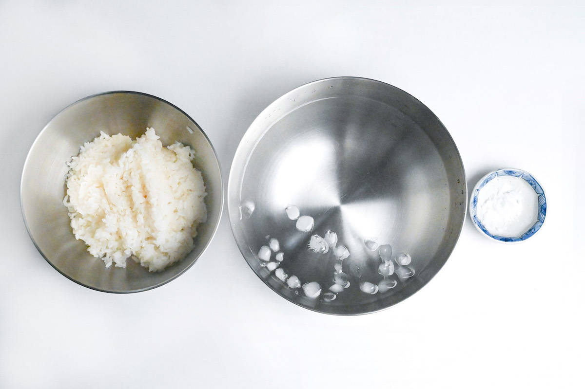 a bowl of cooked white Japanese rice next to a bowl of ice water and a small plate of salt
