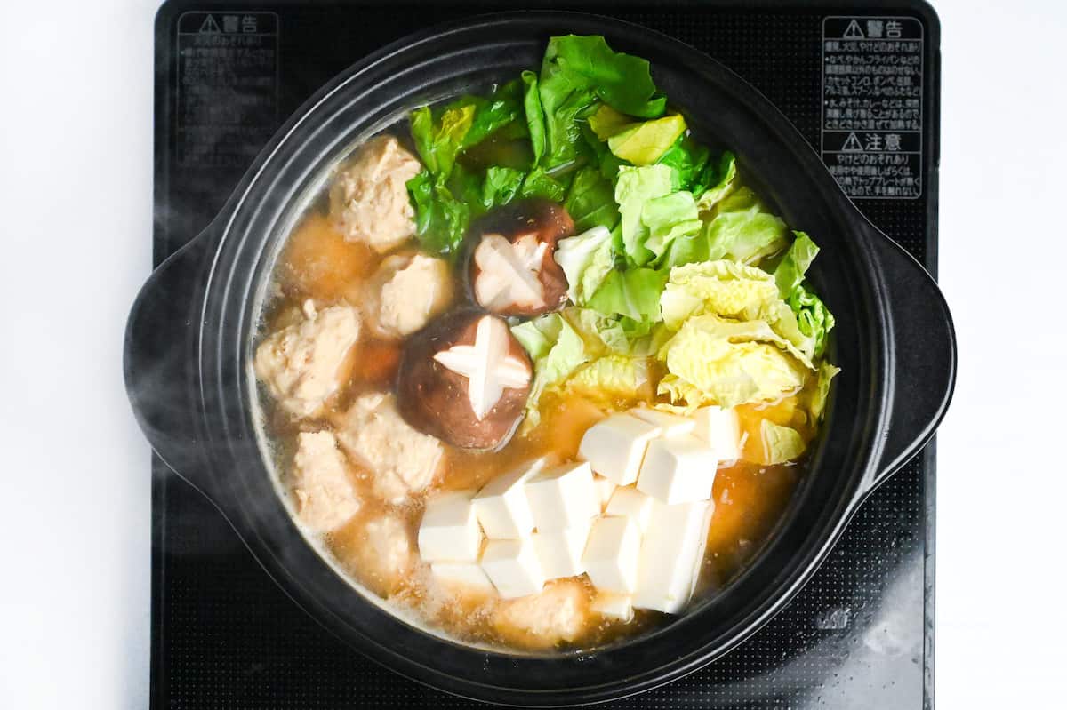 chanko nabe in a black pot on the stove with chicken meatballs, shiitake mushrooms, tofu and naba cabbage