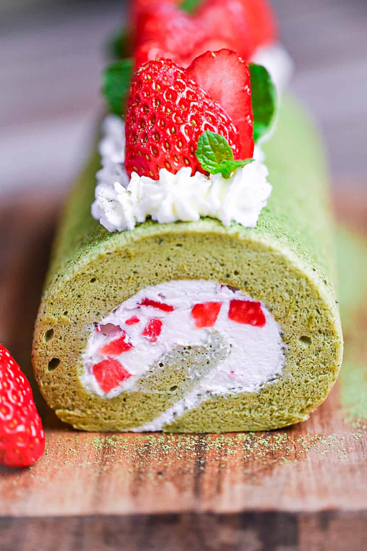 A whole Matcha Roll Cake (Green Tea Swiss Roll) on a wooden chopping board topped with cream, strawberries and mint leaves