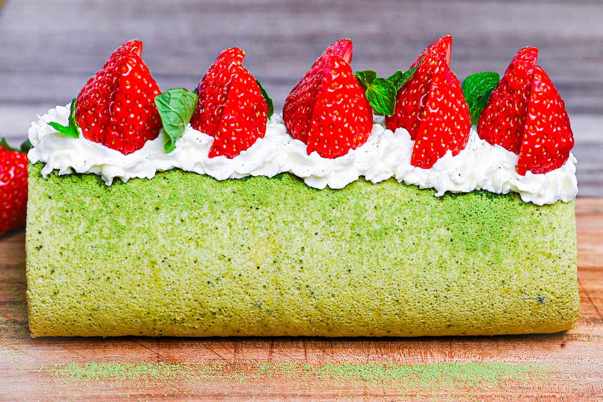 A whole Matcha Roll Cake (Green Tea Swiss Roll) on a wooden chopping board topped with cream, strawberries and mint leaves