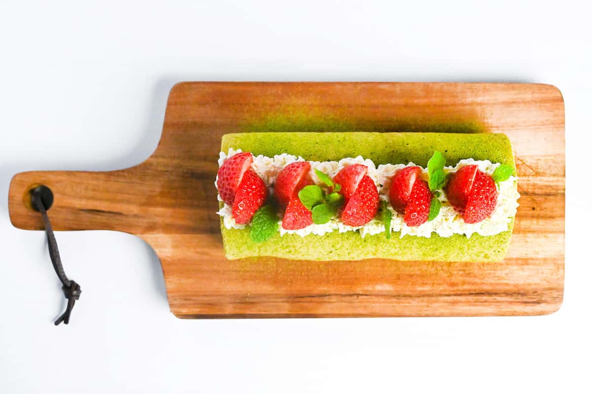 chilled matcha roll cake decorated with a dusting of matcha powder, whipped cream, strawberries and mint leaves