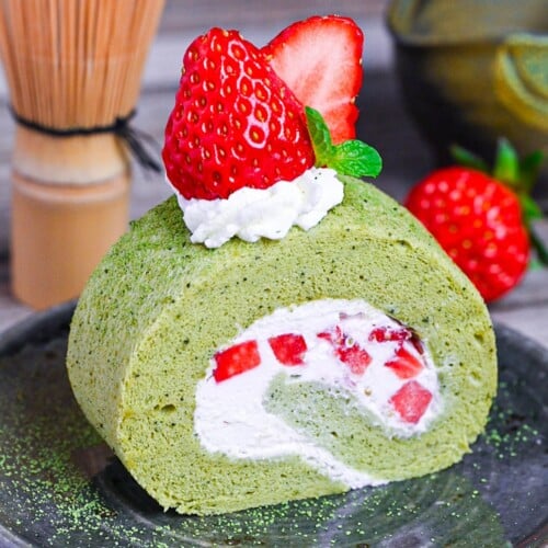 A slice of Matcha Roll Cake (Green Tea Swiss Roll) on a green plate topped with cream, strawberries and a mint leaf