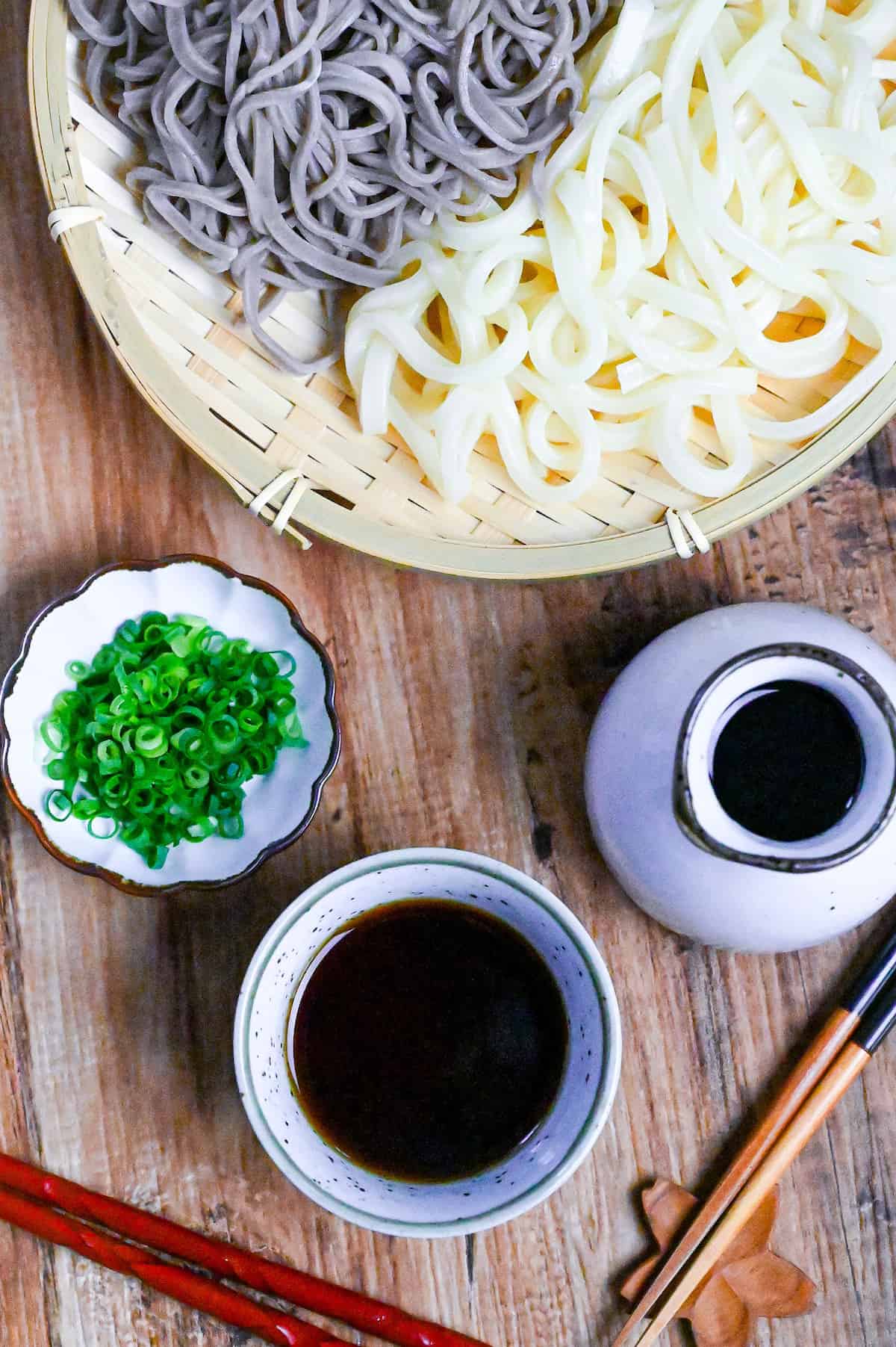 homemade mentsuyu (Japanese noodle dipping sauce) in a white dipping bowl next to a jug of sauce and a tray of udon and soba noodles