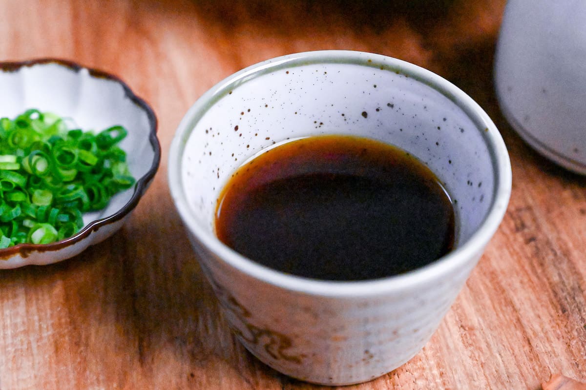 homemade mentsuyu (Japanese noodle dipping sauce) in a white dipping bowl next to a jug of sauce and a tray of udon and soba noodles